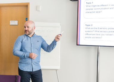 Trainer stands at the front of the room and is pointing towards a presentation displayed on a projector, out of shot. There is a flip chart behind them.