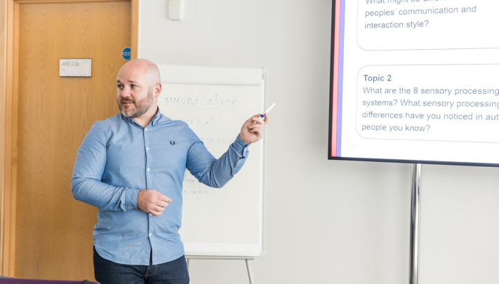 Trainer stands at the front of the room and is pointing towards a presentation displayed on a projector, out of shot. There is a flip chart behind them.