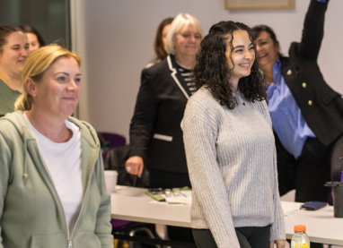 A classroom scene with learners, one standing with raised hand participating in a learning activity whilst others stand facing the front of the room.