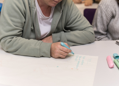 Two people working together at a desk, one writing on paper with a blue marker pen whilst the other looks on.